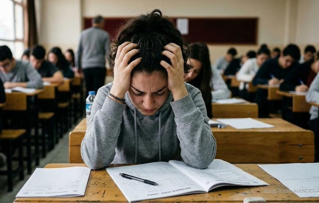 Worried student holding a pen while taking an exam in a classroom with other students in the background.